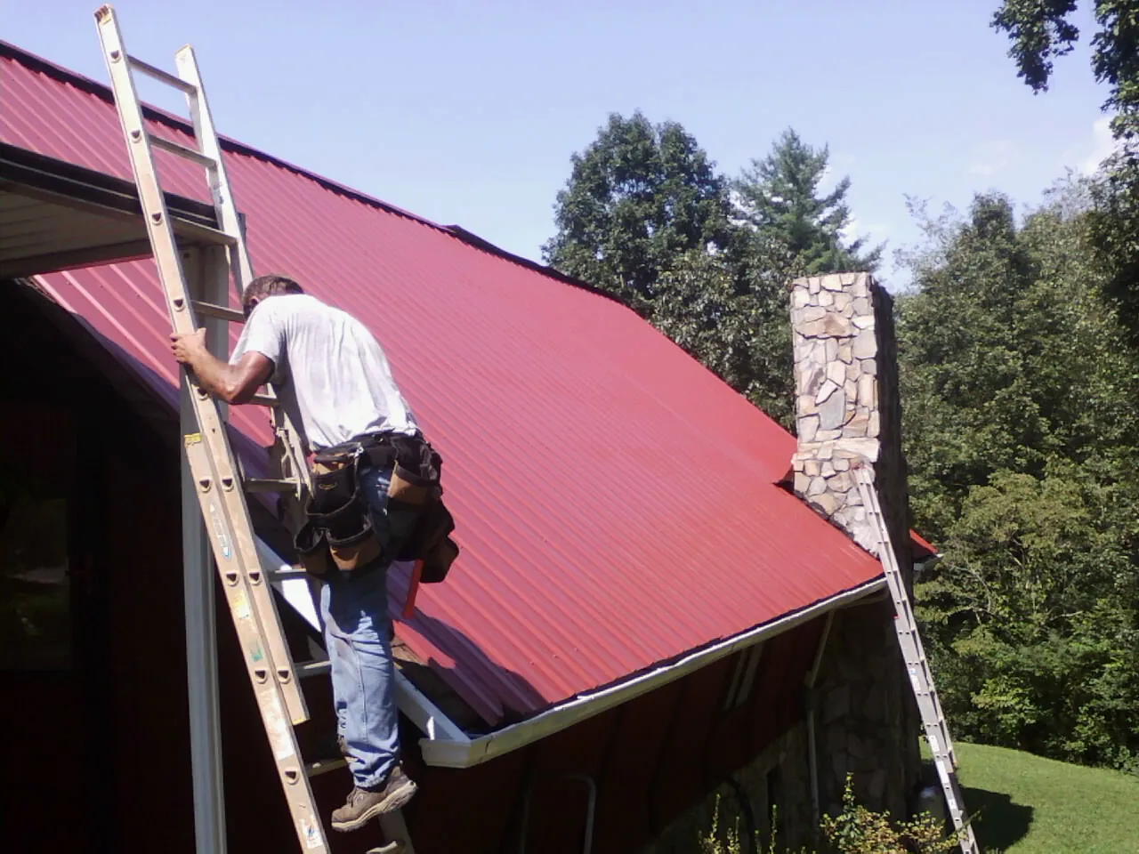 Roofer on a ladder beside a red metal roof and stone chimney — Soffit & Fascia Repair by Roofing Brothers in Harlan